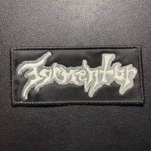 Pode incluir: Um emblema retangular bordado em preto com letras brancas estilizadas que dizem "Igorrr". O emblema tem uma borda preta costurada e uma base de tecido texturizado, exibindo um trabalho de linha intrincado em todo o design.