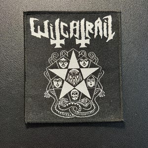 Peut inclure: Un écusson rectangulaire en tissu noir avec des détails brodés en blanc. Le haut affiche le texte WITCHTRAIL dans une police stylisée. En dessous, un pentagramme blanc contient quatre visages et une figure centrale barbue, avec un crâne et deux serpents.