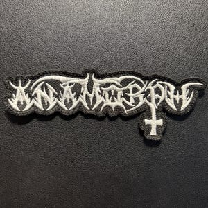 Könnte beinhalten: Ein gestickter Aufnäher mit weißem Garn auf schwarzem Hintergrund. Das Design zeigt den Text ANAMORPH in einer stilisierten, gezackten und scharfen Schriftart. Ein kleines weißes Kreuzsymbol ist im unteren rechten Teil des Schriftzugs integriert.