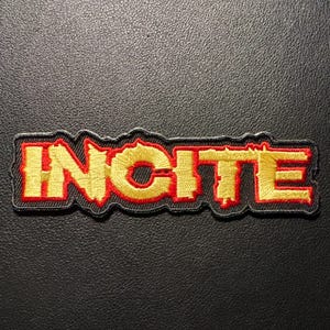 May include: Embroidered patch featuring the word "INCITE" in gold lettering with a red outline. The patch has a black border and is set against a dark background. Suitable for application on clothing or accessories.
