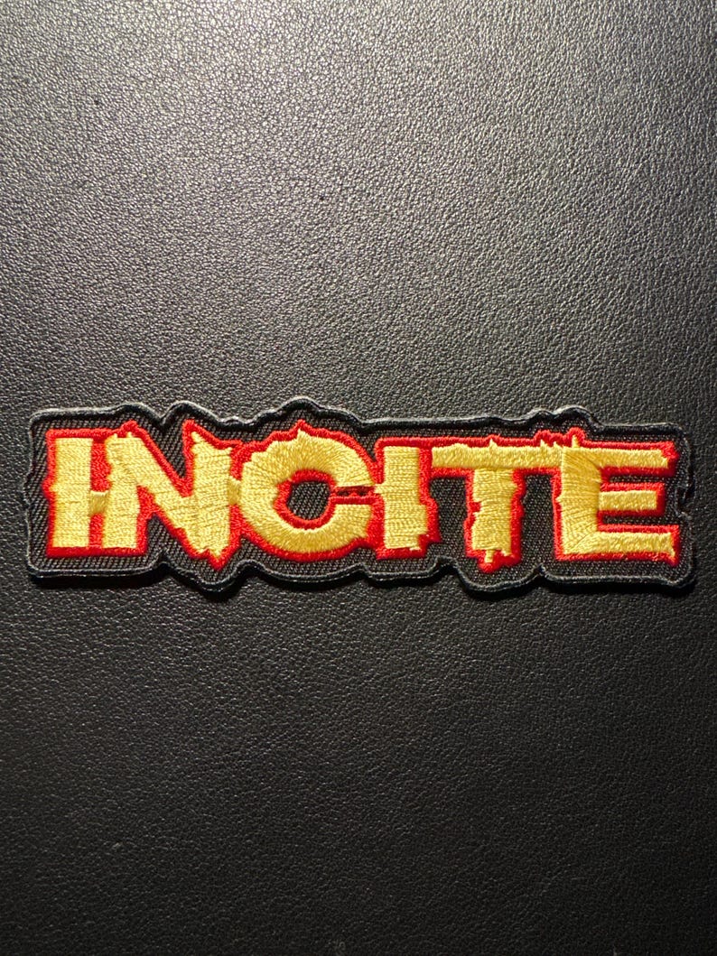 Op de afbeelding: Geborduurde patch met het woord "INCITE" in gouden letters met een rode omlijning. De patch heeft een zwarte rand en is geplaatst tegen een donkere achtergrond. Geschikt voor kleding of accessoires.
