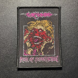 Peut inclure: Un écusson rectangulaire brodé noir avec le nom du groupe Carcass en rose en haut et le titre de l'album Reek of Putrefaction en rose en bas. Le centre présente une illustration graphique d'une personne au visage déformé.