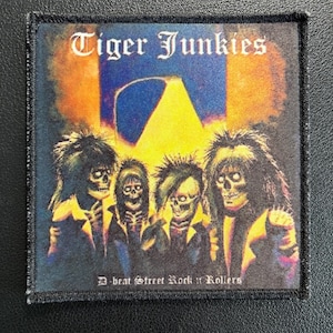 May include: Square patch with the text "Tiger Junkies" in a gothic font. The image features four skeletal figures with long hair, set against a backdrop of orange, yellow, and blue. The text "D-beat Street Rock n Rollers" is at the bottom.