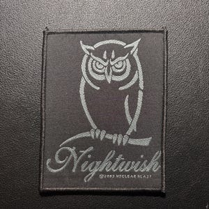 May include: A black square patch featuring a detailed owl illustration in light gray, perched on a branch. The word "Nightwish" is written in a stylized font below the owl. The patch has a stitched border.