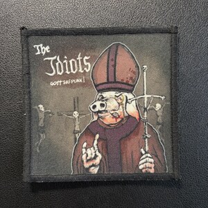 May include: A square fabric patch featuring a graphic illustration of a pig dressed in religious vestments, including a miter and robes. The text reads The Idiots and GOTT SEI PUNK. The pig holds a cross and makes a hand gesture against a dark background.