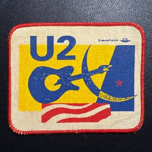 Peut inclure: Écusson rectangulaire avec le logo du groupe U2. Le design comprend une guitare bleue, un croissant de lune et une étoile rouge sur un fond jaune et bleu. Le texte "U2" est présent. Bordure rouge.
