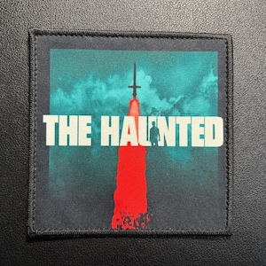 May include: Square patch featuring the text "THE HAUNTED" in white. The design includes a teal and red background with a silhouette of a person and a cross-shaped object.