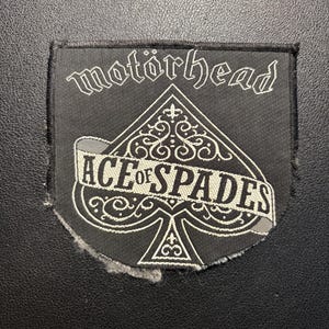 Può includere: Una toppa in tessuto nero con il testo ricamato in bianco "motörhead" nella parte superiore e un grande simbolo di picche al centro. Un nastro orizzontale sopra il picche mostra il testo bianco "ACE OF SPADES" con dettagli in filigrana.