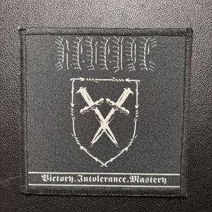 Può includere: Una toppa quadrata in tessuto nero con un disegno ricamato in bianco. La parte superiore presenta un testo stilizzato, il centro mostra uno scudo contenente due spade incrociate e la parte inferiore include il testo Victory. Intolerance. Mastery.