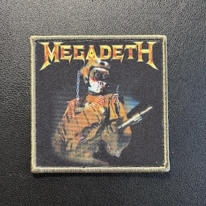 May include: A square fabric patch featuring the Megadeth logo in yellow and orange at the top. The center displays a graphic of a figure in a military-style helmet and uniform, rendered in shades of brown, orange, and black with a stitched border.