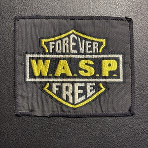 Könnte beinhalten: Ein rechteckiger schwarzer Aufnäher mit den Worten "FOREVER W.A.S.P. FREE" in gelben und weißen Buchstaben. Der Aufnäher hat ein Schild- und Banner-Design mit einem schwarzen Rand.