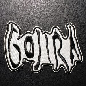 May include: Embroidered patch featuring the word "GOJIRA" in a stylized, dripping font. The patch has a white background with black lettering and a black border. The patch is likely for a band or music merchandise.