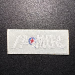 May include: A rectangular white embroidered patch with the word "SUMMA" in white thread. A small circular emblem with red and blue details is centered above the "M". The patch is on a dark textured surface.