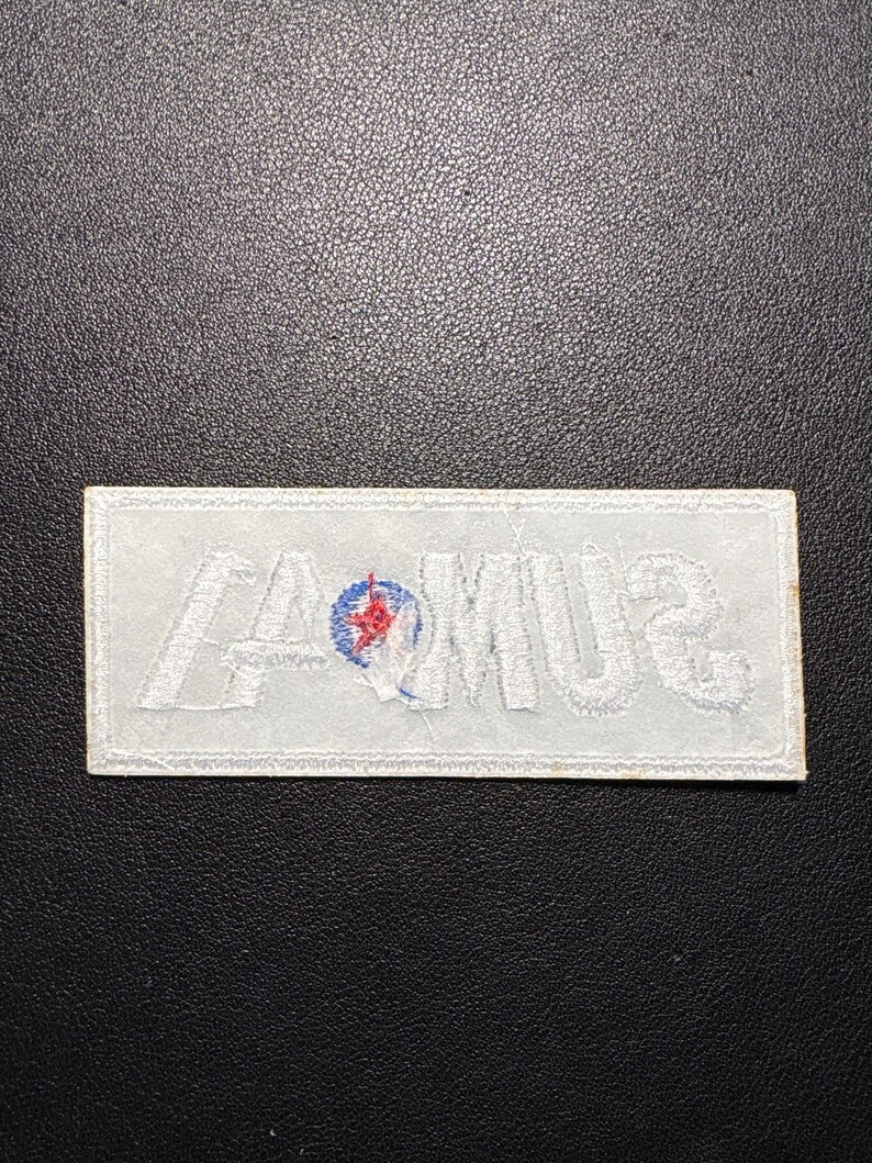 May include: A rectangular white embroidered patch with the word "SUMMA" in white thread. A small circular emblem with red and blue details is centered above the "M". The patch is on a dark textured surface.
