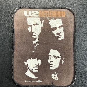 Pode incluir: Um emblema de tecido retangular com fundo castanho escuro apresentando um gráfico a preto e branco dos quatro membros da banda U2. O texto U2 RATTLE AND HUM está impresso a laranja no topo, com texto de direitos de autor na base.