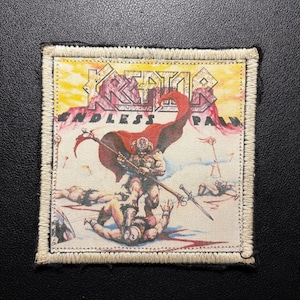 May include: Embroidered patch featuring the band name "Kreator" and the text "Endless Pain" in a stylized font. The patch depicts a warrior scene with a red cape, axes, and fallen figures, with a cream-colored border.