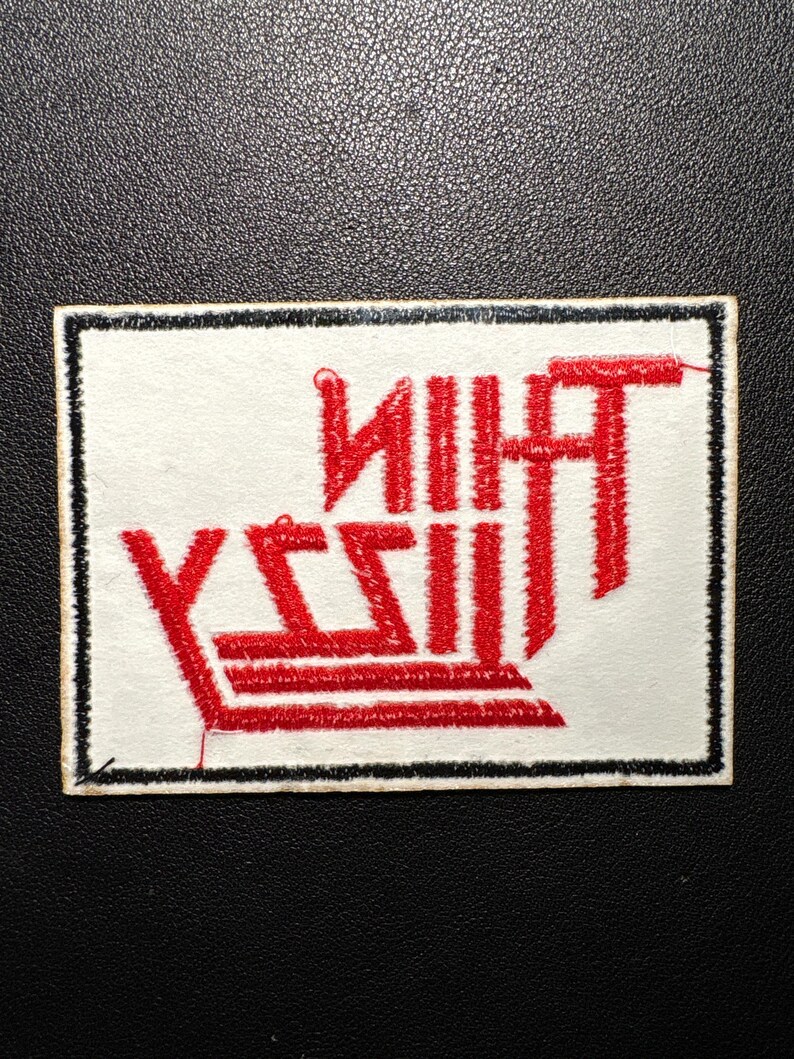 Pode incluir: Patch retangular branco com borda preta, com o texto vermelho "THIN LIZZY". O texto &eacute; estilizado com uma fonte vertical e em negrito. O patch &eacute; provavelmente um emblema bordado ou impresso.