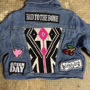 Pode incluir: Uma jaqueta jeans azul com um painel central geométrico em preto e branco com detalhes em rosa. Os patches incluem o texto BAD TO THE BONE, GREEN DAY e BLINK-182, além de um coração rosa atravessado por uma flecha e duas cerejas.