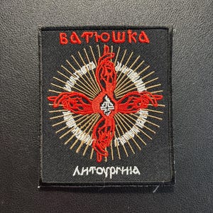 Könnte beinhalten: Ein rechteckiger, schwarzer Stickaufnäher mit einem zentralen, roten, kreuzartigen Design aus stilisierten Händen. Über dem Design steht der rote Text "ВАТЮШКА" und darunter der weiße Text "ЛИТУРГІЯ". Das Motiv ist von feinen goldenen und weißen Strahlen umgeben.