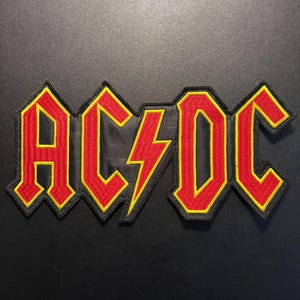 Op de afbeelding: Een geborduurde stoffen patch met het AC/DC-logo. De letters zijn rood met een gele rand en een gele bliksemschicht scheidt de AC van de DC. Het hele ontwerp is omlijnd met zwart garen, wat zorgt voor een opvallende en krachtige uitstraling.