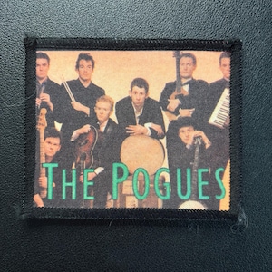 Könnte beinhalten: Rechteckiger Aufnäher mit einem Bandfoto von The Pogues. Das Bild zeigt eine Gruppe von Musikern in Anzügen, die Instrumente halten. Der Bandname "THE POGUES" ist in grünen Blockbuchstaben auf der Unterseite des Aufnähers aufgedruckt.