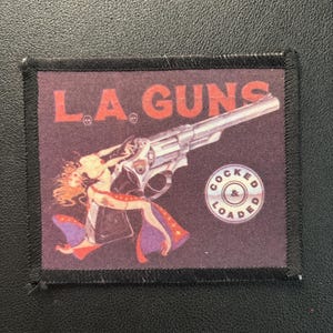 Könnte beinhalten: Ein rechteckiger Aufnäher mit dem Text "L.A. GUNS" in Rot, über einer großen silbernen Revolver und einer Pin-up-Girl-Illustration. Der Aufnäher zeigt auch den Text "COCKED & LOADED" in einem kreisförmigen Design.