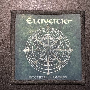 Könnte beinhalten: Quadratischer Aufnäher mit dem Wort "Eluveitie" in einer stilisierten Schriftart oben. Der Aufnäher zeigt ein weißes kreisförmiges Design mit Text und einer zentralen Figur auf einem dunkelgrünen Hintergrund. Der Text "Evocation II - Pantheon" steht unten.