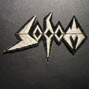 Könnte beinhalten: Gestickter Aufnäher mit dem Bandnamen "Sodom" in einem geometrischen Design. Der Aufnäher hat einen schwarzen Hintergrund mit dem Bandnamen in Schwarz und Off-White. Der Aufnäher ist wahrscheinlich für eine Jacke oder eine Tasche.