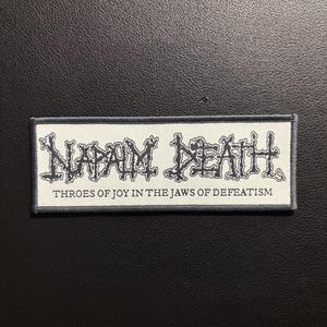 May include: A rectangular embroidered patch with a white background and a dark gray border. The patch features the text NAPALM DEATH in a stylized black font, with the text THROES OF JOY IN THE JAWS OF DEFEATISM printed in a smaller black font below.