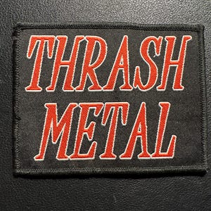 Könnte beinhalten: Schwarzer rechteckiger Aufnäher mit dem Schriftzug "THRASH METAL" in Rot mit weißer Umrandung. Der Aufnäher hat einen schwarzen Rand und ist zum Aufnähen auf Kleidung oder Accessoires gedacht.