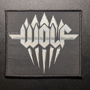 Op de afbeelding: Een zwarte rechthoekige patch met het woord "WOLF" in zilver, gestileerd met scherpe, hoekige lettertypen en een gekarteld ontwerp. De patch heeft een zwarte rand en de tekst "©2009 WOLF" is zichtbaar.