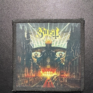 Könnte beinhalten: Quadratischer Aufnäher mit dem Bandnamen "Ghost" in goldfarbenen Buchstaben über einer detaillierten Illustration einer gotischen Stadtlandschaft mit einer schädelartigen Struktur. Der Aufnäher hat einen schwarzen Rand und einen dunklen Hintergrund.