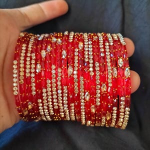 May include: A collection of red bangles, each embellished with lines of small, clear and gold-coloured gemstones. The bangles are held in a hand against a dark backdrop, highlighting their detailed design and vivid colour.