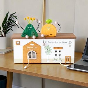 Puede incluir: Un organizador de cables de madera blanco y marrón con forma de casa, con el texto "Baking Hut" y "Dis Bonjour Avec Un Gâteau". Dos figuras de madera con brazos y piernas están sentadas en el tejado. Un portátil y una planta están en el fondo.