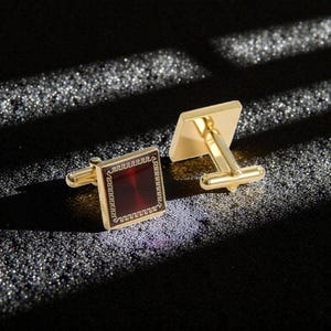 Op de afbeelding: Goudkleurige manchetknopen met een vierkant, dieprood middenstuk en een decoratieve rand. De manchetknopen hebben een klassiek ontwerp, perfect voor formele gelegenheden. De achtergrond is donker en gestructureerd.