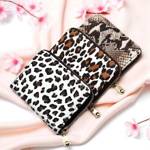 Puede incluir: Tres carteras cuadradas con cremallera y diseños de animales. Las carteras presentan un fondo blanco con manchas de leopardo marrón, un estampado de leopardo marrón y beige, y un estampado de piel de serpiente. Cada cartera tiene una cremallera dorada.