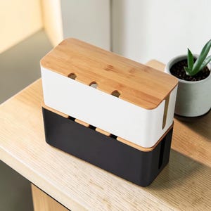 May include: Two cable management boxes, one black and one white, stacked on top of each other. Each box has a rectangular shape with a bamboo-colored wooden lid. The lids have three small openings. A small potted plant is in the background.