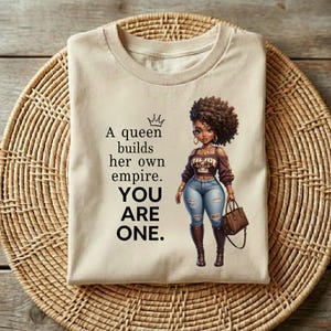 May include: Beige t-shirt with the text "A queen builds her own empire. YOU ARE ONE." and an illustration of a woman with curly hair, wearing a brown top, jeans, and boots. The woman is holding a brown purse.