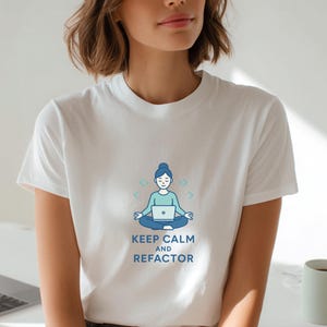 Könnte beinhalten: Weißes T-Shirt mit einer blau-türkisen Grafik einer Person, die mit einem Laptop meditiert, darunter der Text "KEEP CALM AND REFACTOR". Das T-Shirt wird von einer Person in einem hellen Raum getragen.