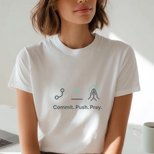 Könnte beinhalten: Weißes T-Shirt mit dem Text "Commit. Push. Pray." und drei minimalistischen grafischen Symbolen. Das Shirt besteht aus einem weichen Material und wird getragen. Ein Laptop und eine Tasse sind im Hintergrund zu sehen.