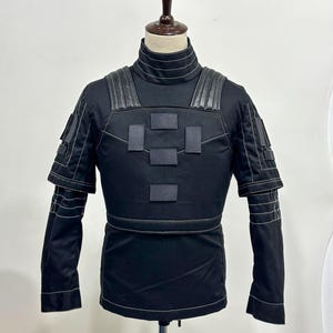 Puede incluir: Chaqueta t&aacute;ctica negra con cuello alto y paneles reforzados en hombros y pecho. La chaqueta presenta m&uacute;ltiples parches rectangulares y detalles de costura blancos. Las mangas tienen pu&ntilde;os en capas. La chaqueta se exhibe en un maniqu&iacute;.