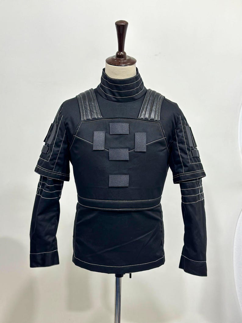 Puede incluir: Chaqueta t&aacute;ctica negra con cuello alto y paneles reforzados en hombros y pecho. La chaqueta presenta m&uacute;ltiples parches rectangulares y detalles de costura blancos. Las mangas tienen pu&ntilde;os en capas. La chaqueta se exhibe en un maniqu&iacute;.