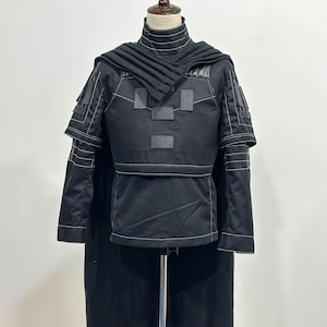Puede incluir: Traje negro con cuello alto, detalles en los hombros y capa con capucha. El atuendo presenta detalles de costura blancos y parches rectangulares en el pecho. El disfraz se exhibe en un maniqu&iacute;.