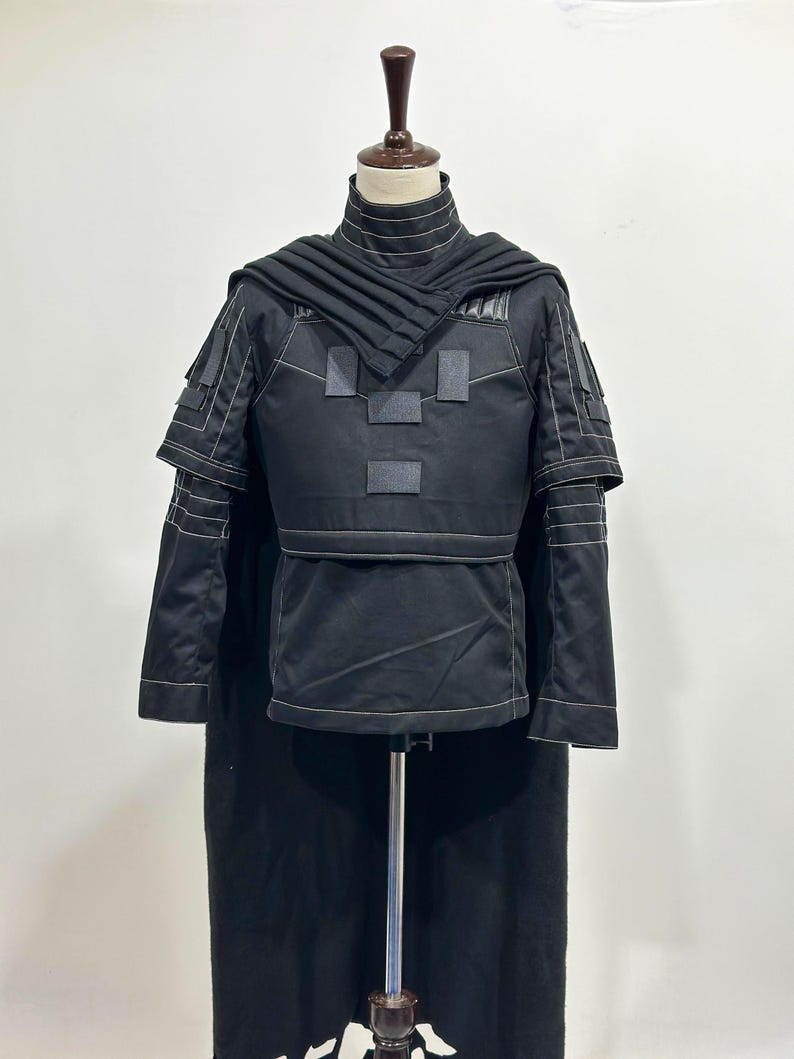 Puede incluir: Traje negro con cuello alto, detalles en los hombros y capa con capucha. El atuendo presenta detalles de costura blancos y parches rectangulares en el pecho. El disfraz se exhibe en un maniqu&iacute;.