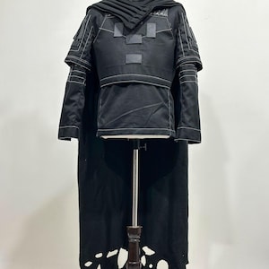 Puede incluir: Traje negro con cuello alto, detalles en los hombros y una larga capa. El atuendo presenta parches rectangulares y detalles de costura blancos. La capa tiene un dise&ntilde;o &uacute;nico e irregular. El disfraz se exhibe en un maniqu&iacute;.