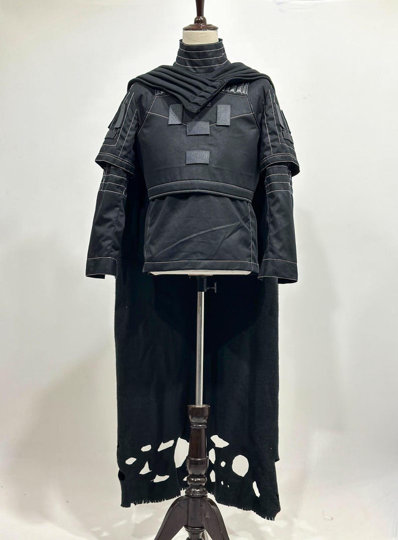 Puede incluir: Traje negro con cuello alto, detalles en los hombros y una larga capa. El atuendo presenta parches rectangulares y detalles de costura blancos. La capa tiene un dise&ntilde;o &uacute;nico e irregular. El disfraz se exhibe en un maniqu&iacute;.