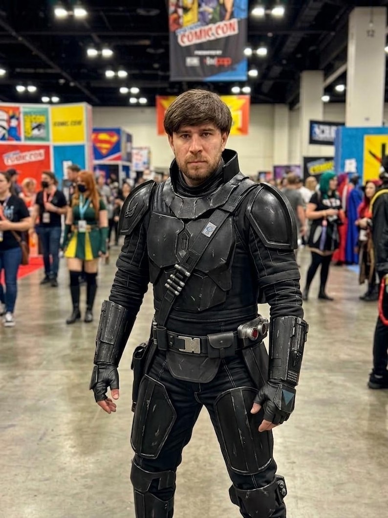 Puede incluir: Una persona con un disfraz negro con armadura, guantes y cintur&oacute;n utilitario, posando en un evento de Comic-Con. El disfraz tiene un dise&ntilde;o futurista con m&uacute;ltiples paneles y detalles. El fondo muestra a otros asistentes y pancartas del evento.