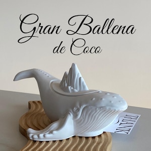 Puede incluir: Un objeto decorativo blanco con forma de ballena con una estructura similar a una montaña en su parte posterior, sobre una base de madera. La ballena tiene detalles y está sobre una superficie clara. El texto "Gran Ballena de Coco" está arriba.