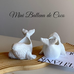 Puede incluir: Dos macetas blancas con forma de ballena sobre una bandeja de madera. Las macetas tienen texturas detalladas y están en diferentes poses. El texto "Mini Ballena de Coco" está arriba.