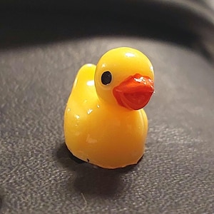 May include: A close-up of a yellow duck shoe charm with an orange beak and black eyes. The charm is on a black shoe. A yellow sunflower shoe charm is visible on the left side of the image.